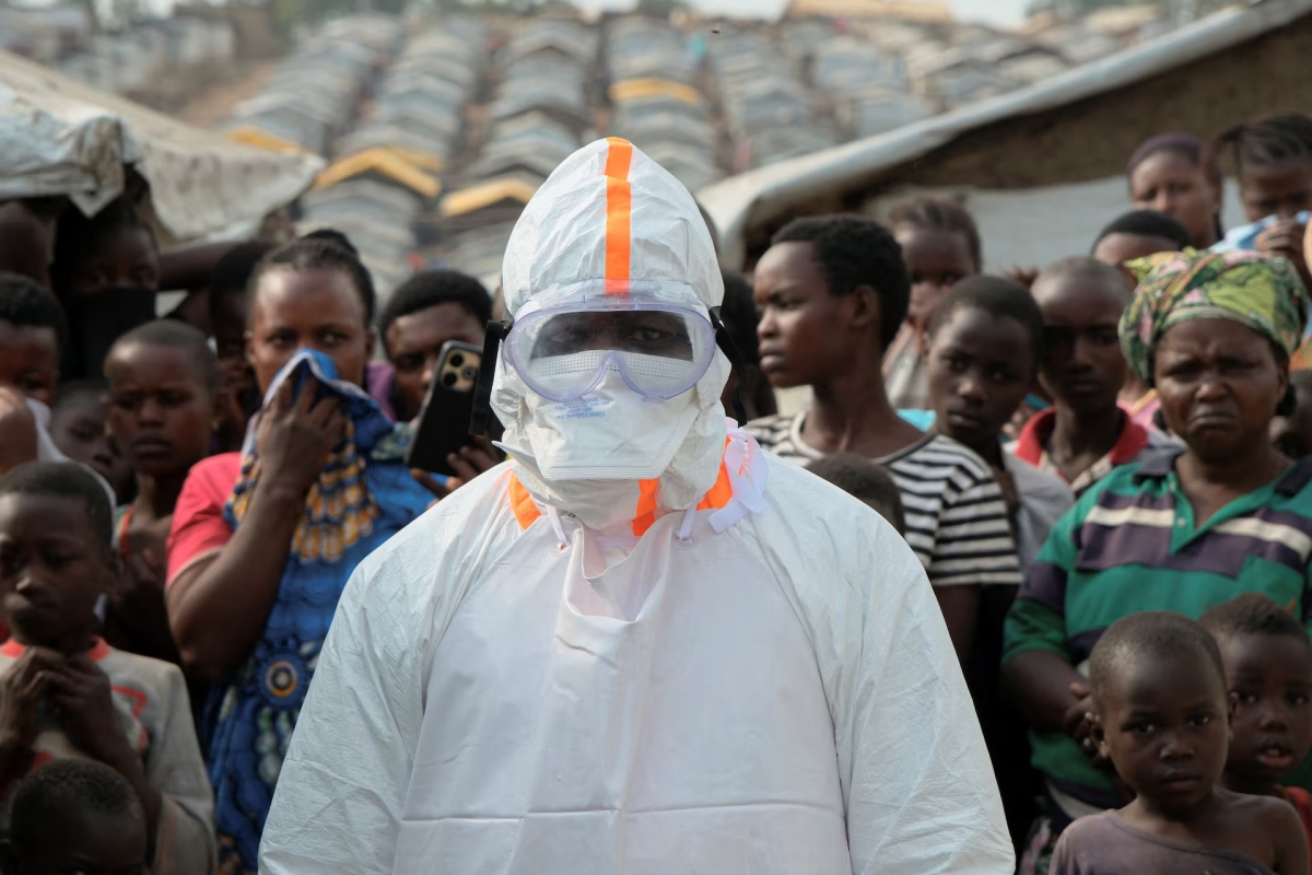 A health worker in personal protective equipment (PPE) stands near displaced people waiting for the burial of Ebola victims at the Kigonze displaced persons camp, one month after an outbreak was declared, in Bunia, eastern Democratic Republic of Congo, June 18, 2026. (REUTERS/Gradel Muyisa Mumbere)